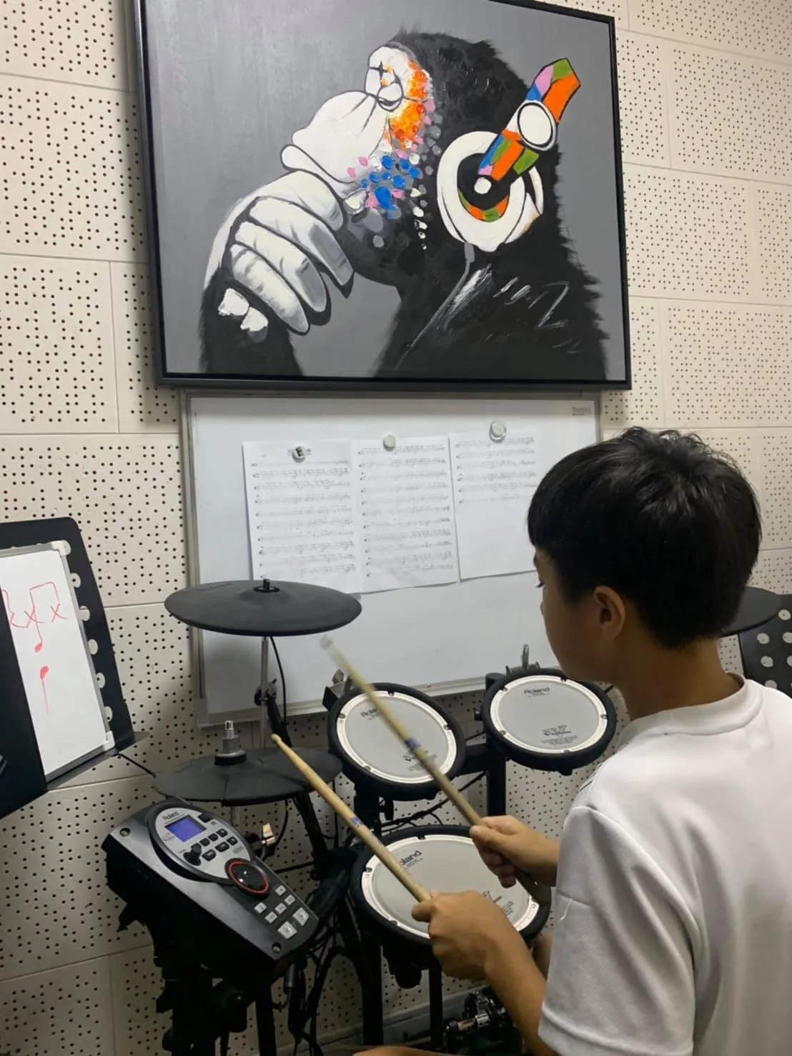 爵士鼓教室裡,學生背對鏡頭練習電子鼓,牆上掛著戴耳機的猩猩畫作