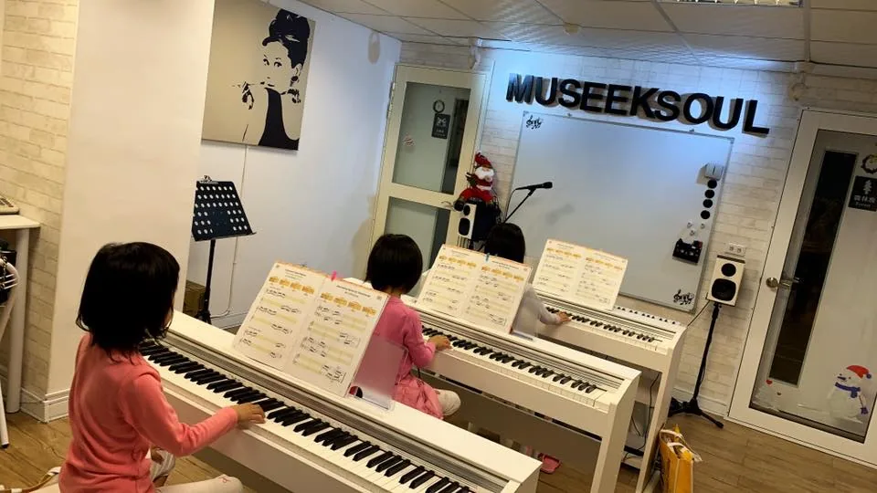 團班教室裡,三位孩子並排在白色電鋼琴前練習,白磚牆上是 MUSEEKSOUL 立體字